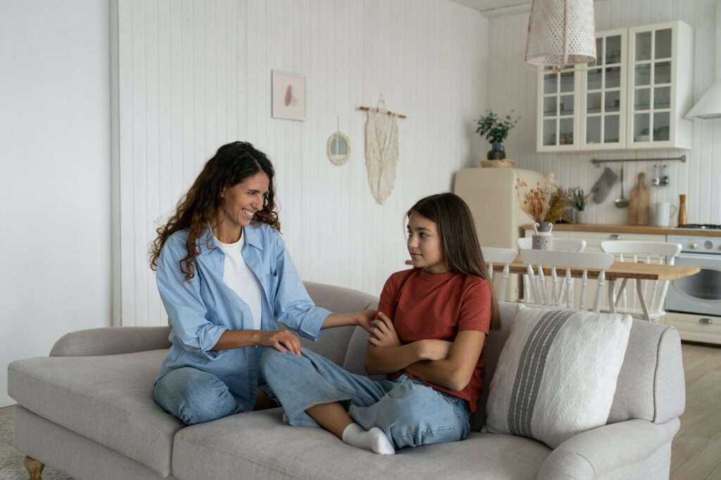 Shy introverted teenage girl sits in closed position on couch with mother wanting to play together