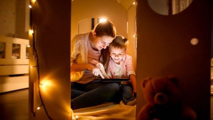 Happy laughing boy with young mother playing games and using tablet computer at night before going