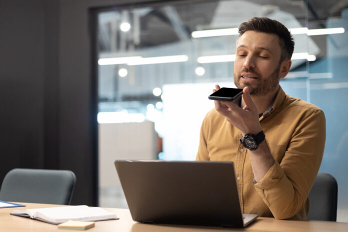 Businessman Using Voice Search On Cellphone Working On Laptop Indoor