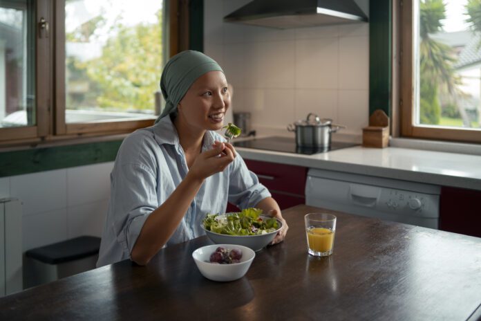The image shows a person sitting at a kitchen table, eating a salad. They are wearing a headscarf and a light blue shirt, and they appear to be smiling while holding a fork with a piece of lettuce. On the table in front of them is a bowl of salad, a small bowl of grapes, and a glass of orange juice. The setting looks like a bright, cozy kitchen with large windows letting in natural light, and there are kitchen appliances like a stove and a pot visible in the background.
