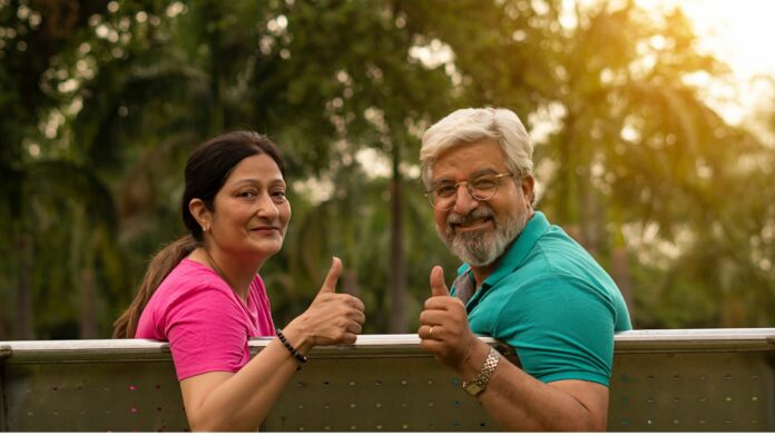 Two smiling senior citizens standing outdoors in a green park-like setting, giving thumbs up while leaning on a railing during warm evening sunlight.